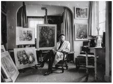 zittend met bril en baret in schildersjas op een stoel in woonkamer tussen zijn werken